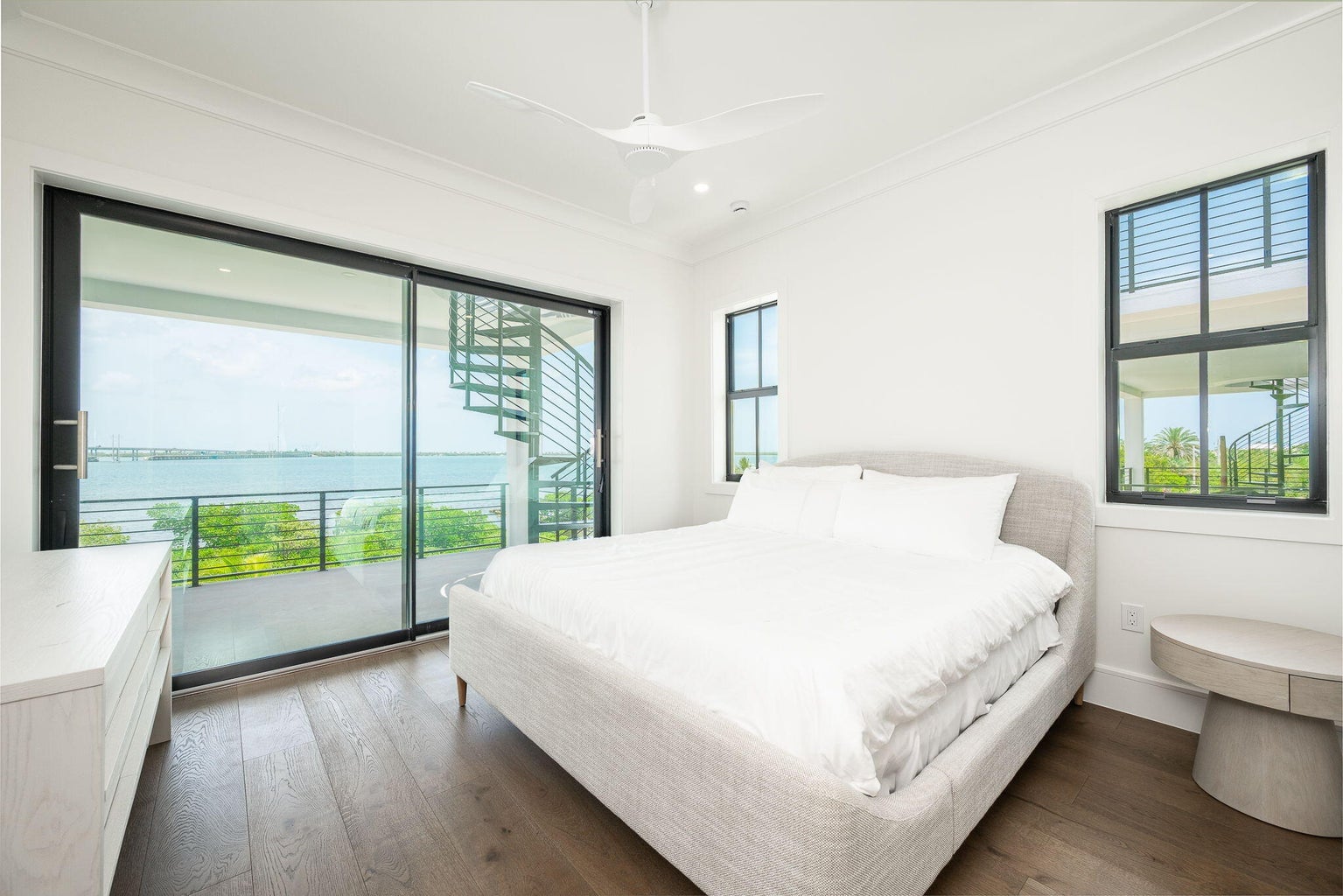 Bedroom with a white upholstered bed and sliding glass doors to a balcony with a spiral stair and water view