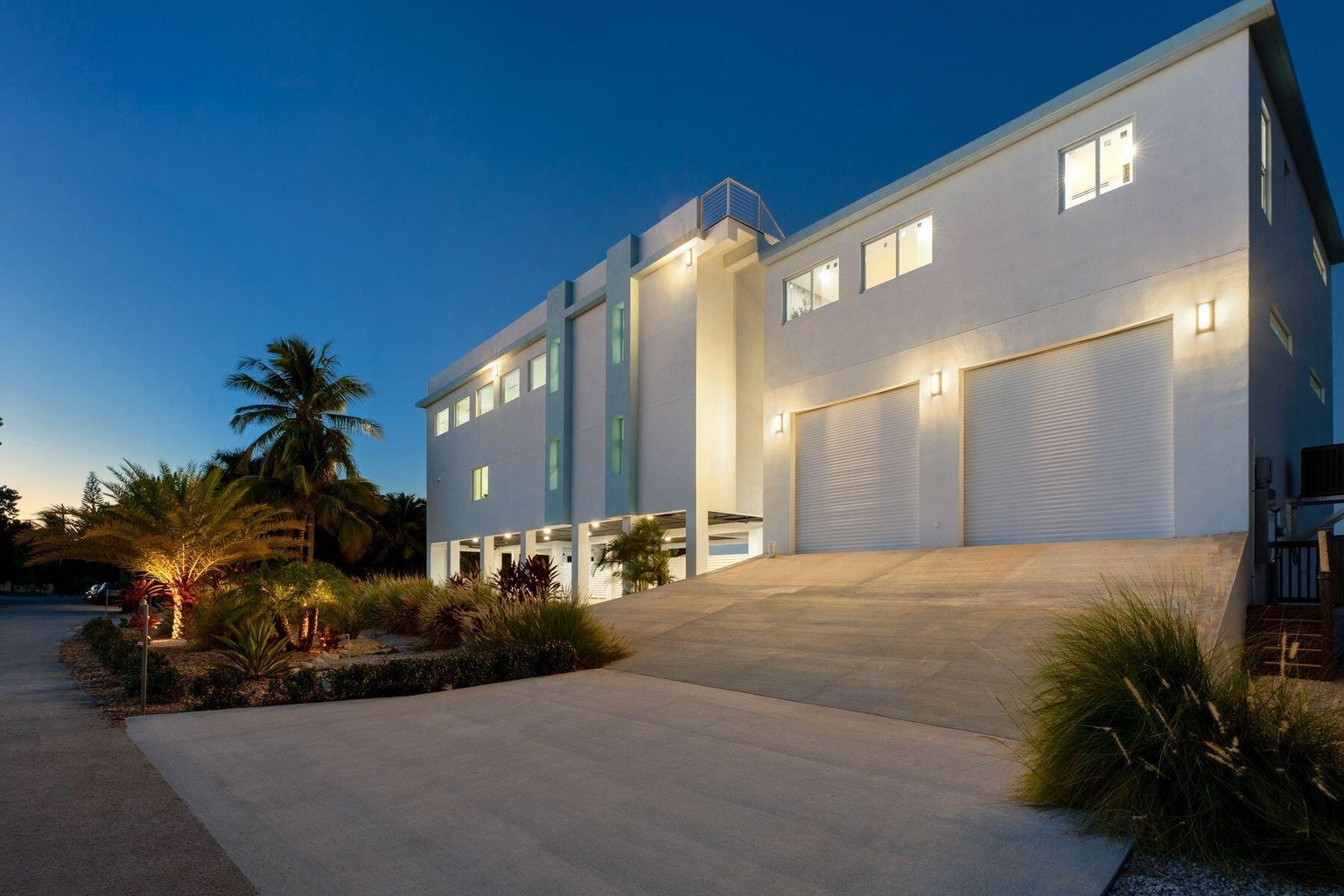 Street side of the residence at twilight with a wide driveway, two garage doors and palms