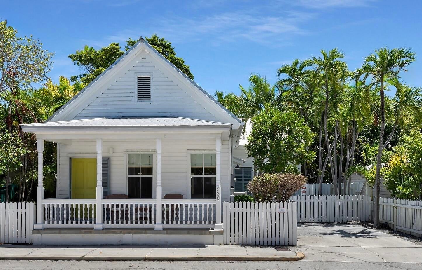 White Conch cottage with a yellow front door, a columned front porch and a white picket fence beneath palms