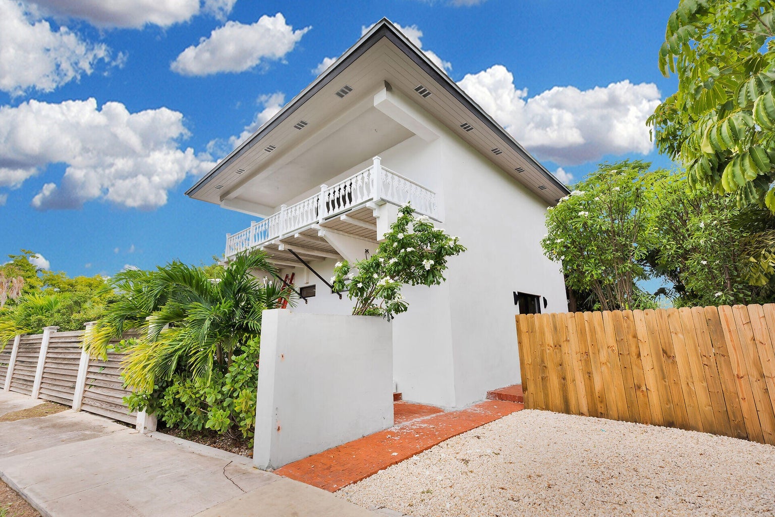 Two-story white home with a covered upper porch and white railings, tropical plantings and a wood fence