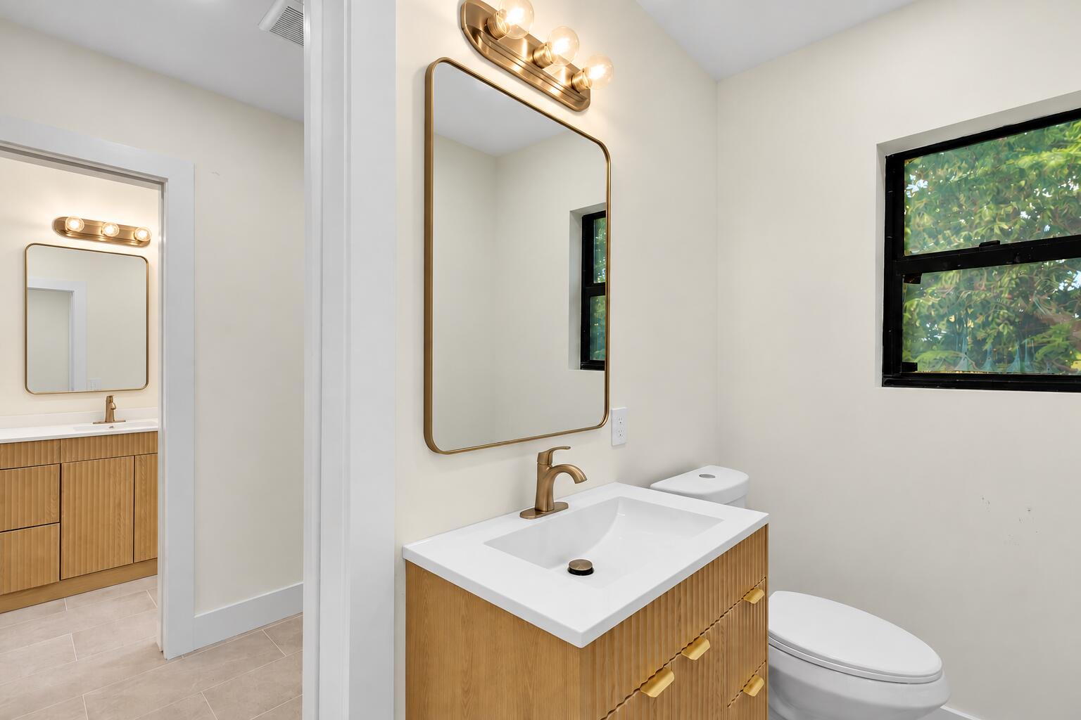 Half bath with a fluted wood vanity, brass mirror and a window onto greenery