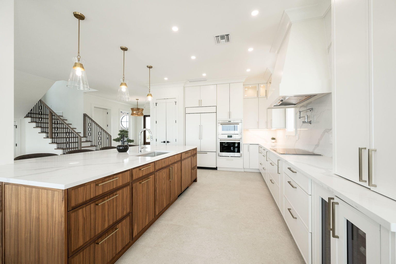 Long walnut island beside a wall of white cabinets and built-in appliances, the staircase beyond