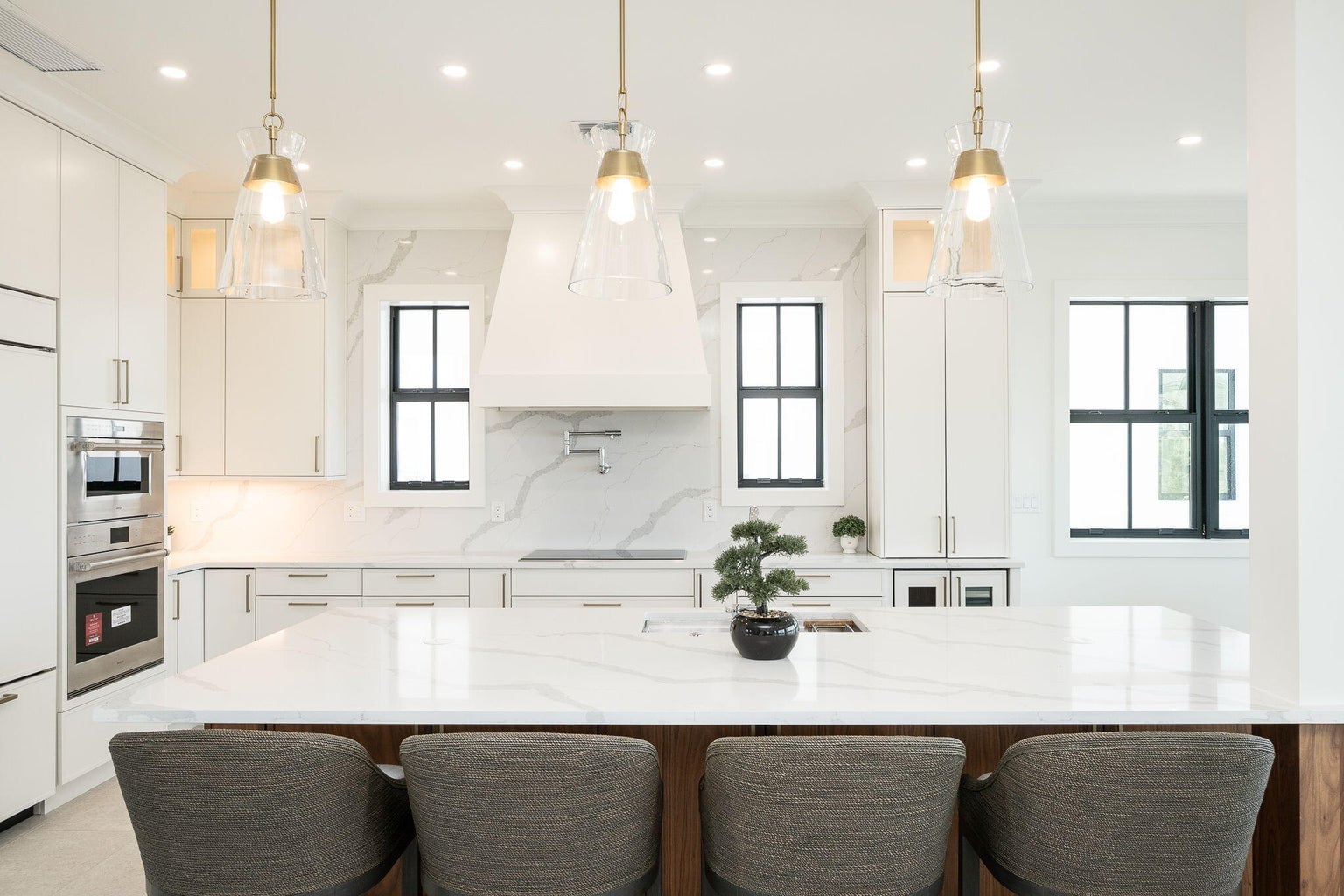 Marble-look island with four gray stools, glass pendants, a white range hood and wall ovens