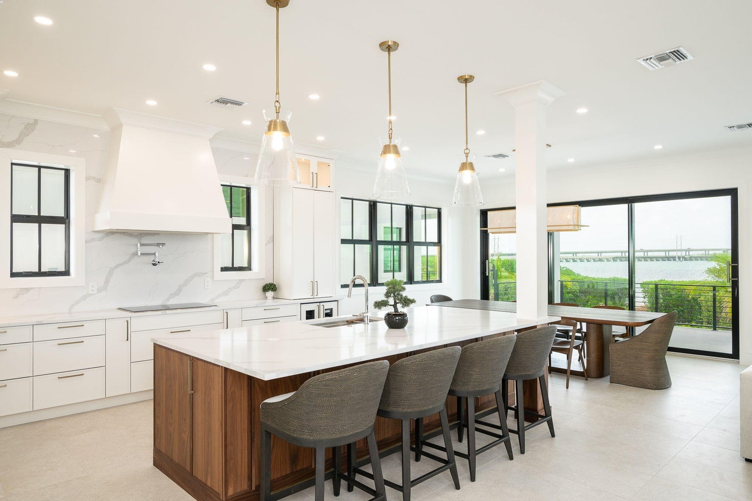 Kitchen with a walnut island and gray stools, white cabinetry, glass pendants and sliding doors to a water view