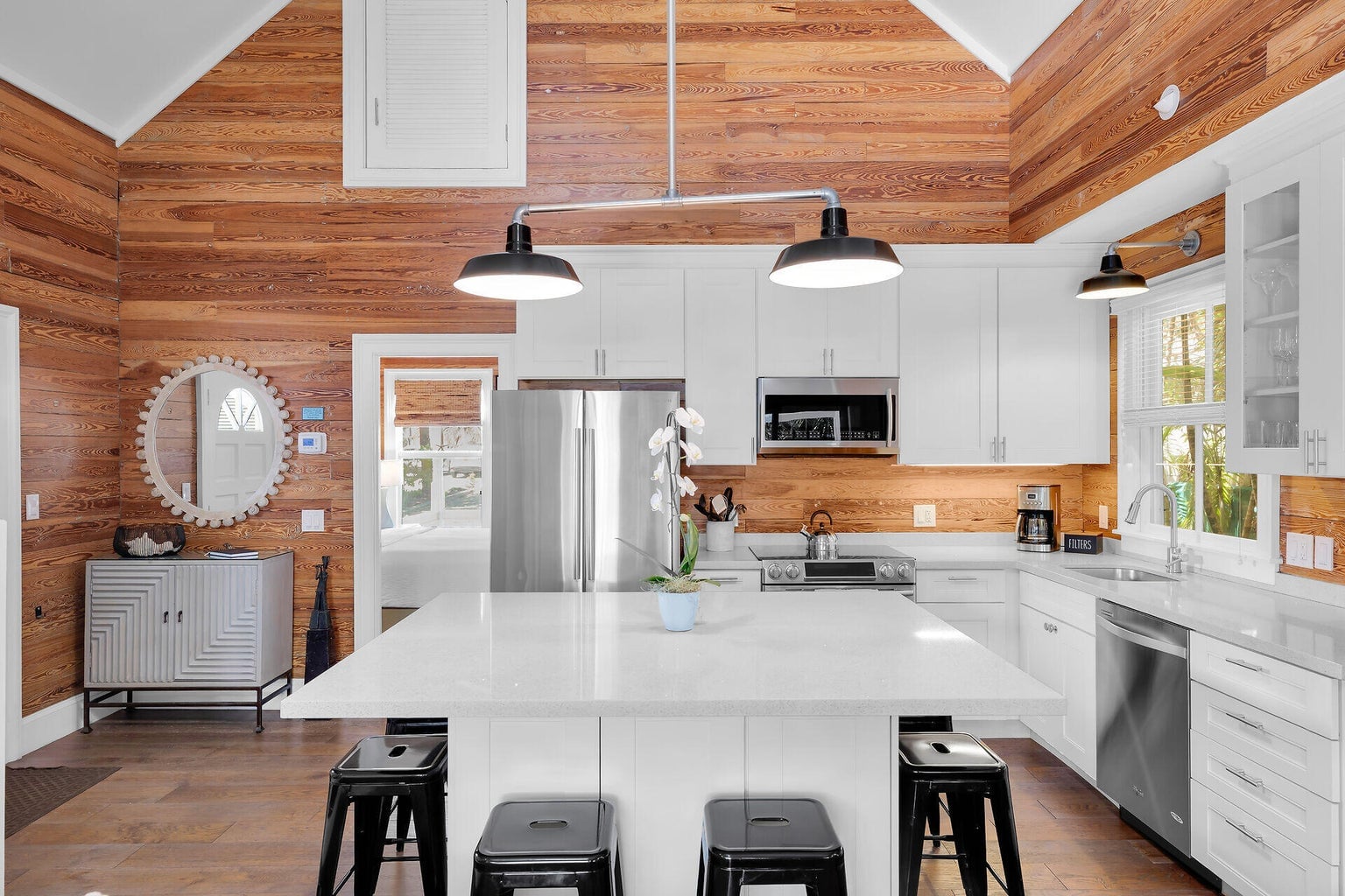 White kitchen with a large quartz island, black stools, stainless appliances and pine-paneled walls