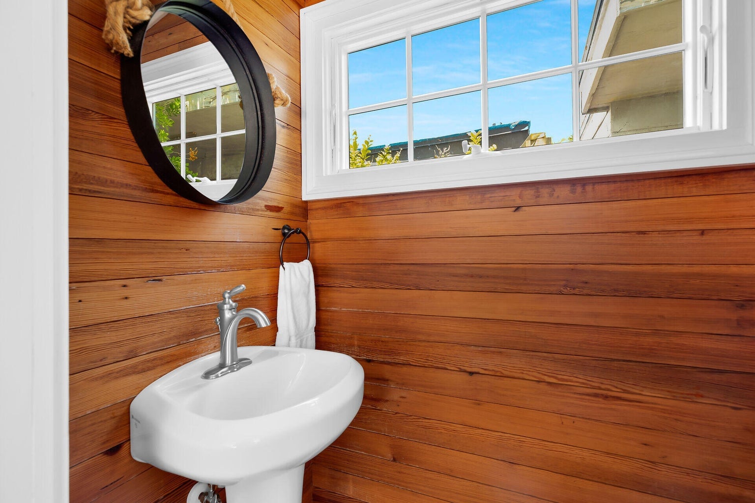 Pedestal sink beneath a round black mirror and a wide window, with pine-paneled walls