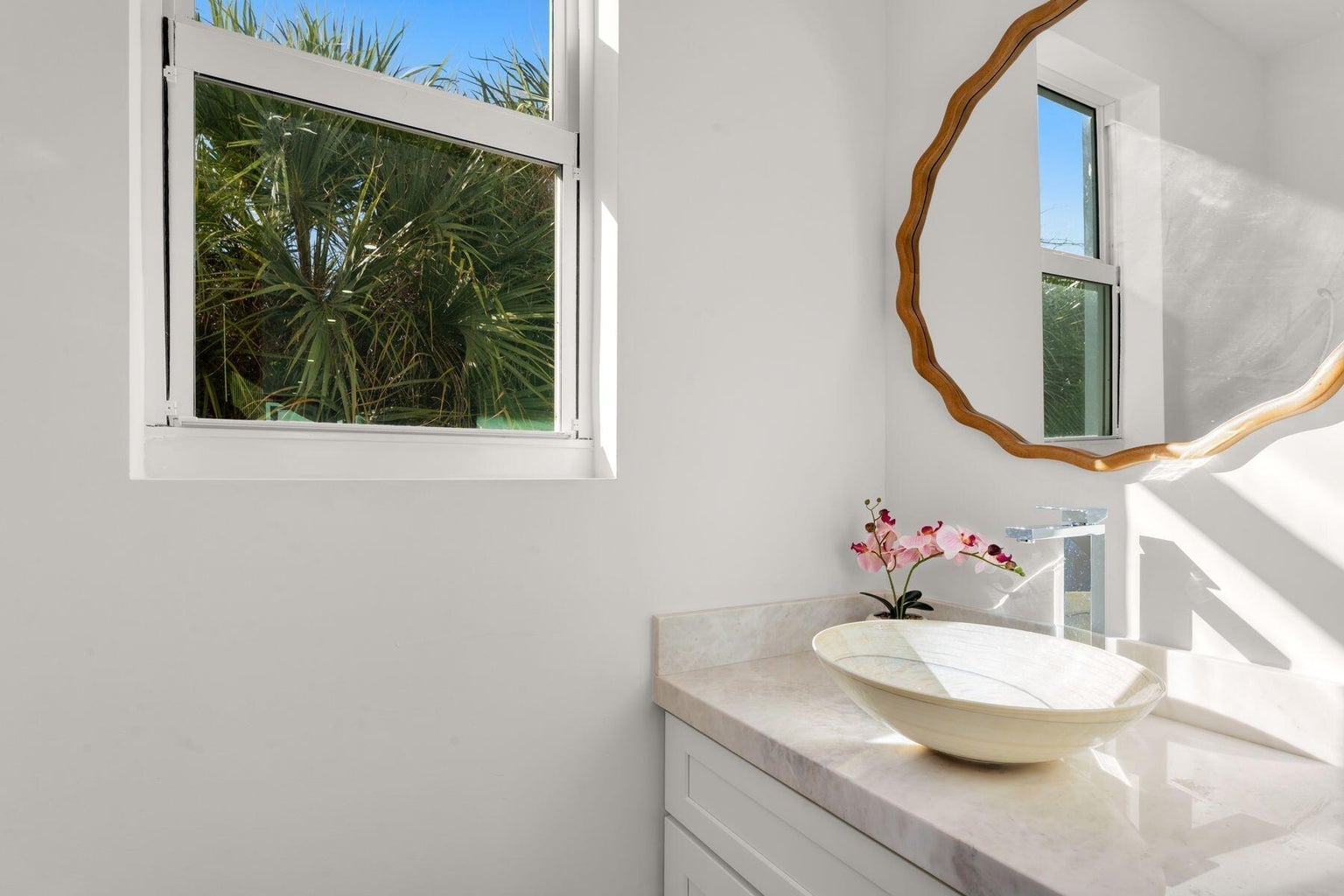 Bathroom vanity with a stone vessel sink, wood-framed mirror and a window onto palms