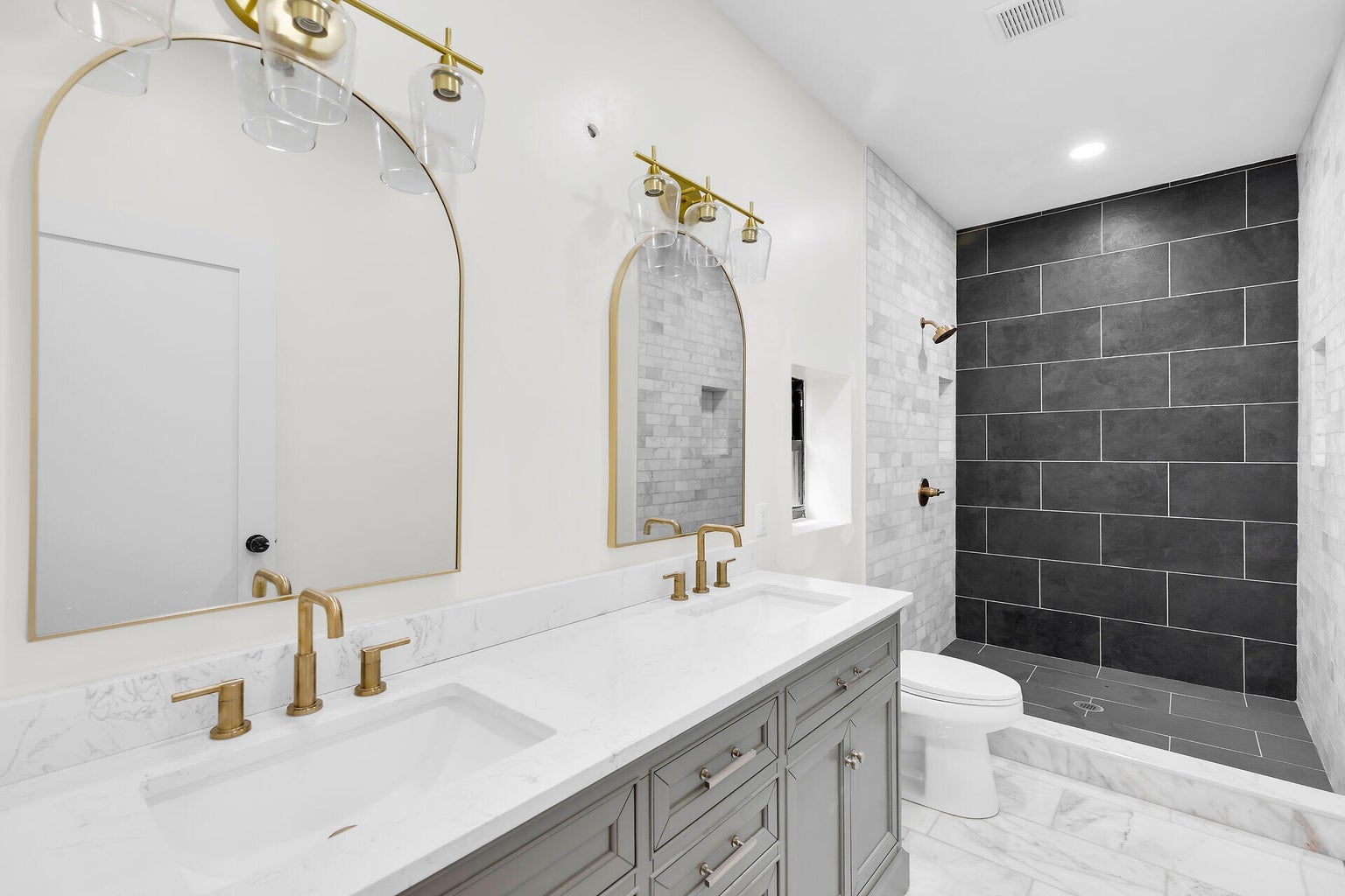 Bathroom with a double vanity, arched brass mirrors and a walk-in shower in dark tile