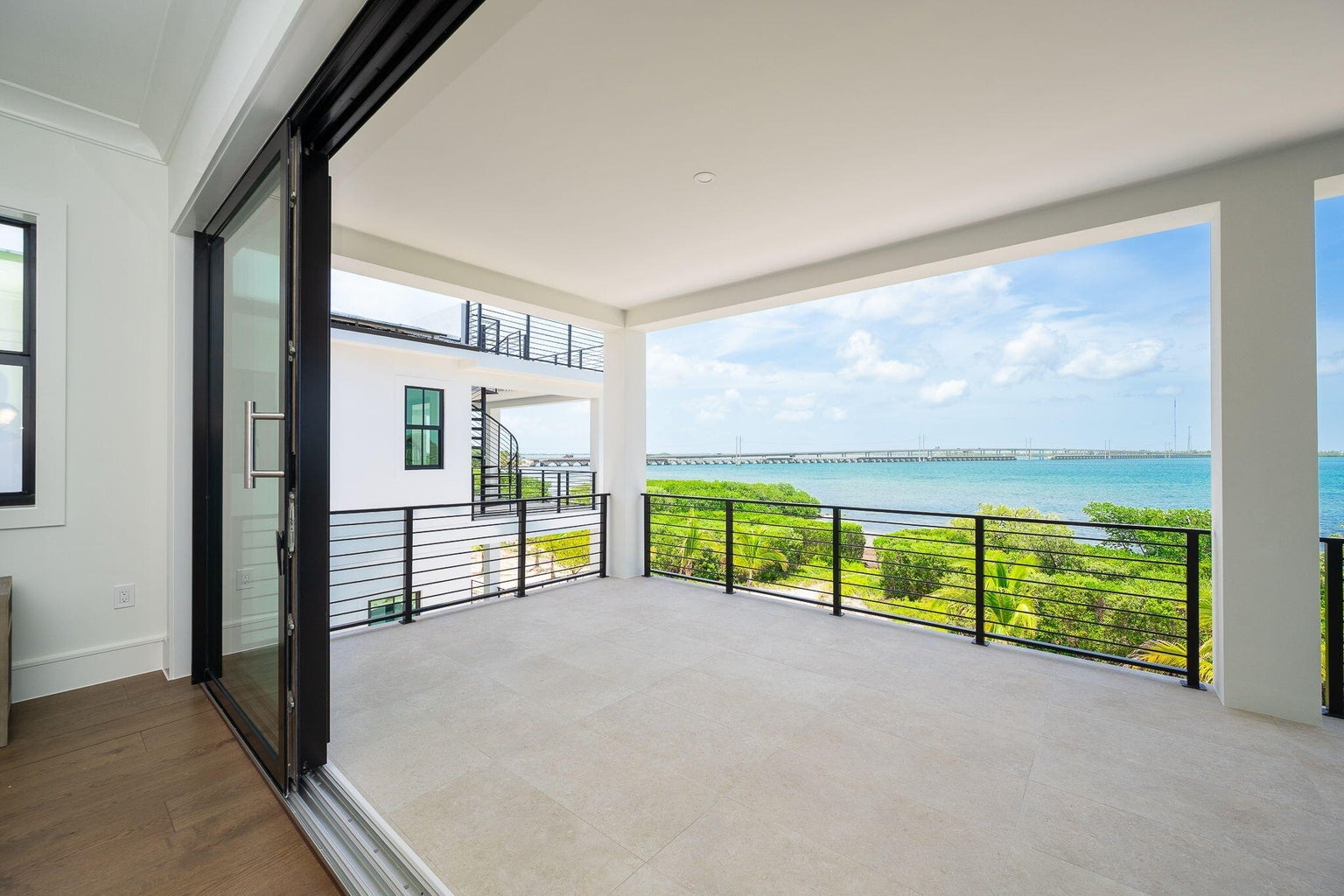 Covered veranda off a bedroom with a black horizontal railing and turquoise water and a long bridge beyond