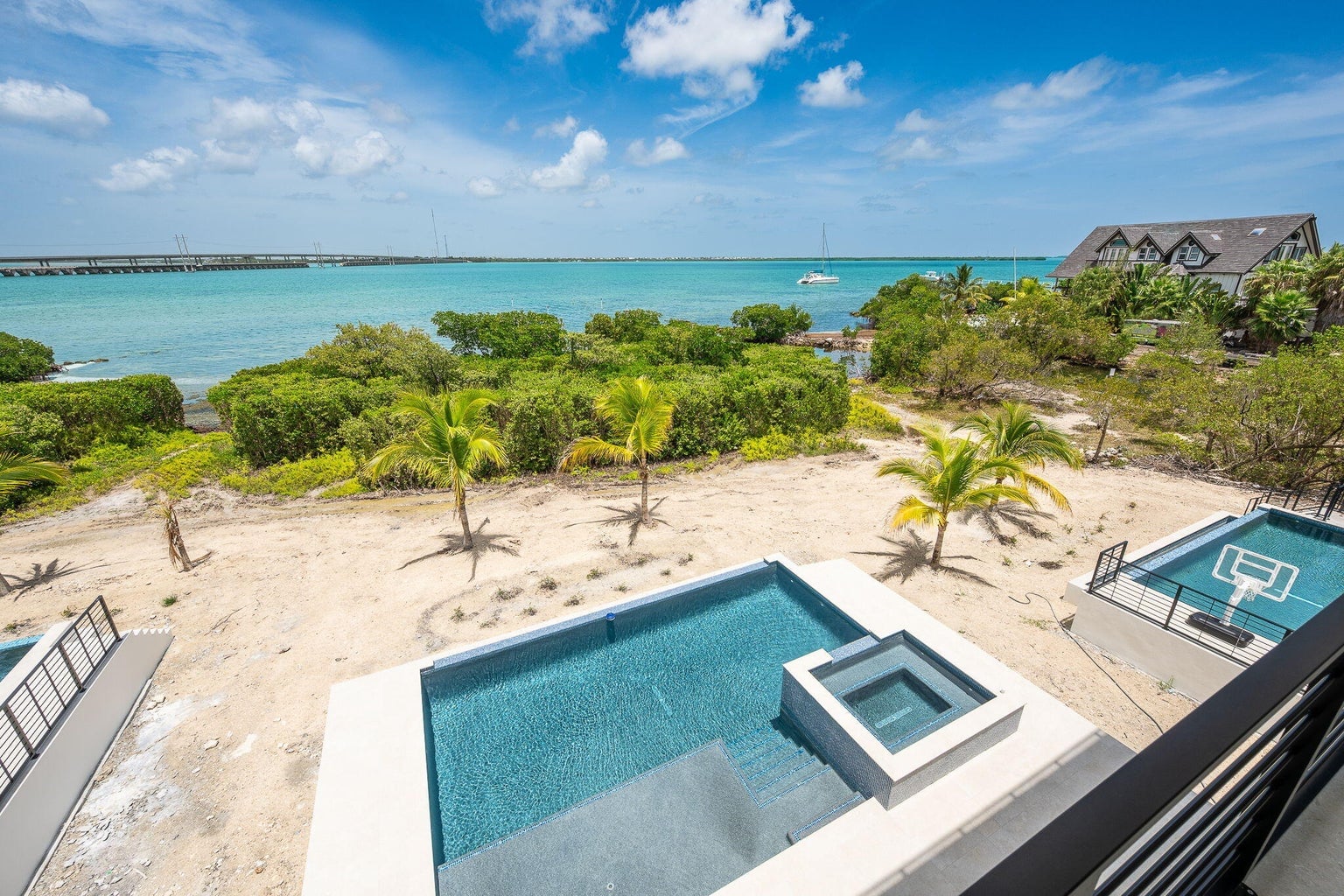 View down from the rooftop to a pool and spa, palms on the sand and turquoise open water with a long bridge on the horizon