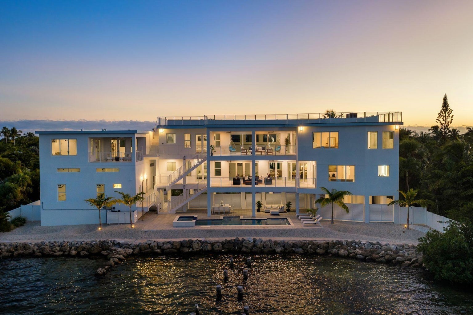 Three-story white residence at dusk seen from the water, with a stone seawall, pool and lit terraces