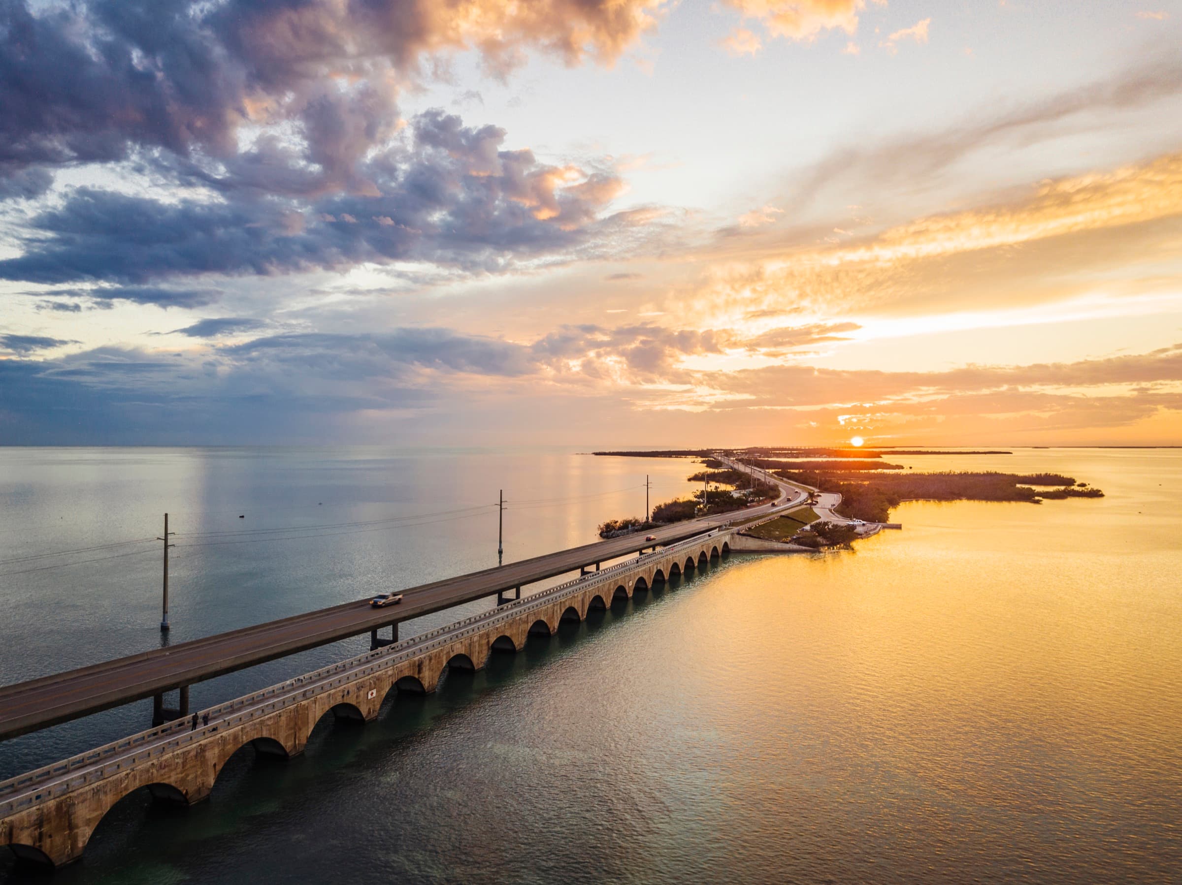 Aerial view at sunset of the Overseas Highway running beside the historic arched railroad bridge across calm water in the Florida Keys.