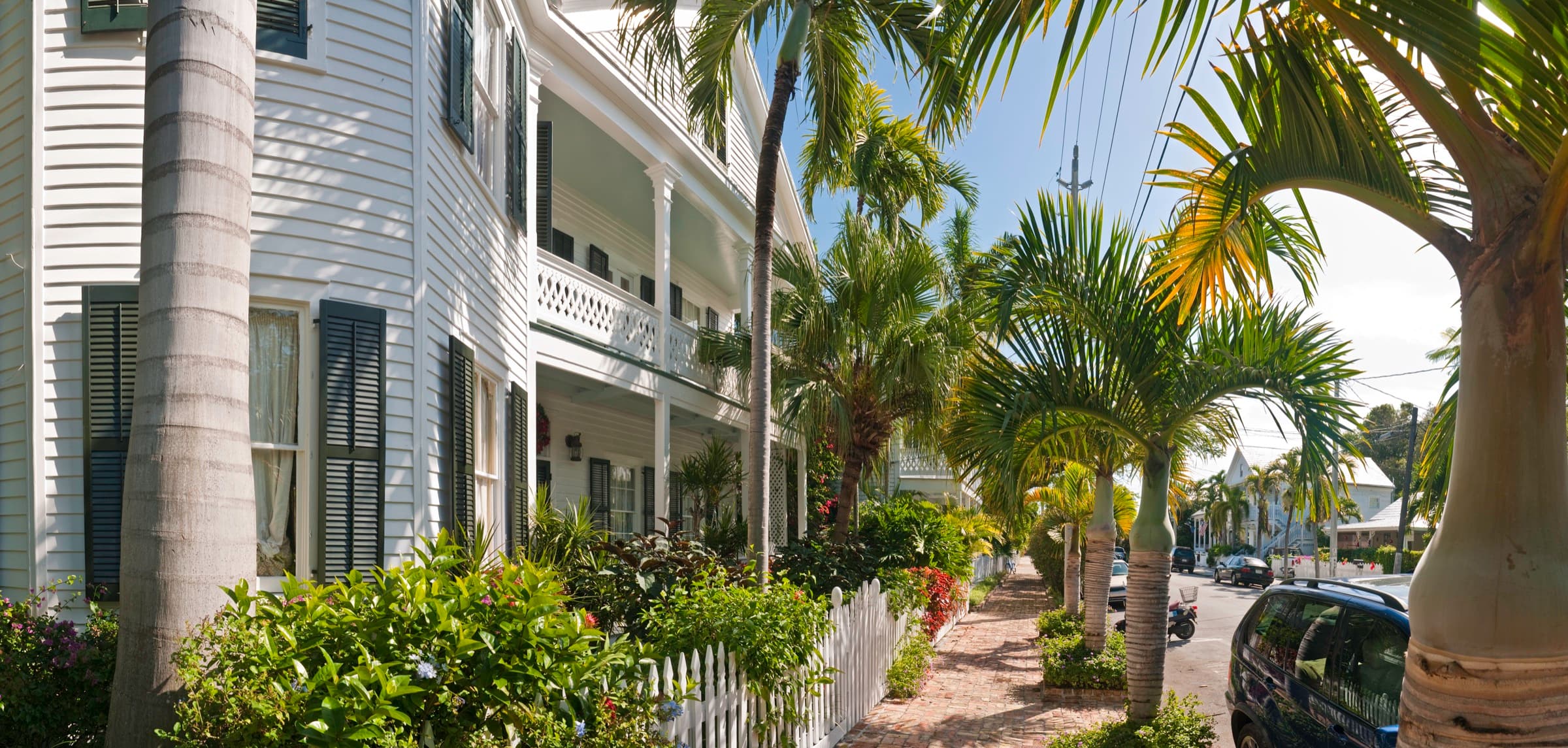 A white two-story Key West house with dark shutters, a wide porch and a white picket fence along a brick sidewalk lined with palm trees.